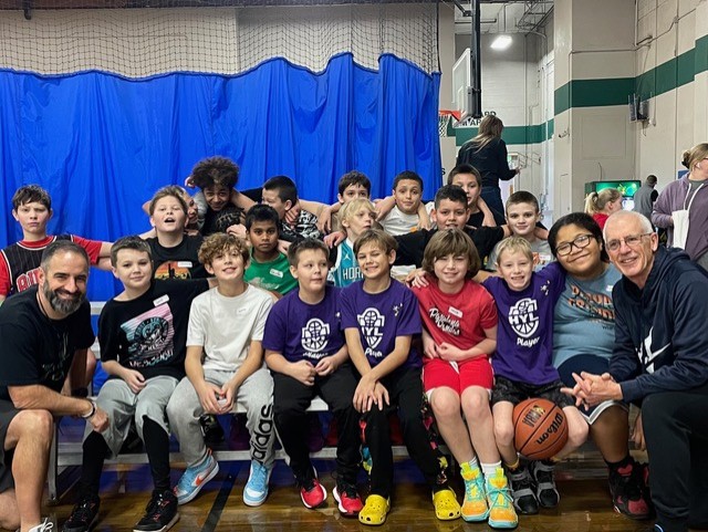 Friends of Rogers basketball team posed on gym bleachers. NBC Camps vice president John Fazio on the team's left and Engagement Champion Doug Jordan on the team's right.